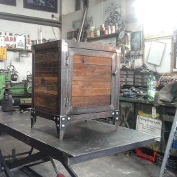 Alfa Welder - Metalowa szafka z drewnianymi drzwiczkami, ustawiona na metalowym stole w warsztacie, widoczne narzędzia i maszyny w tle.