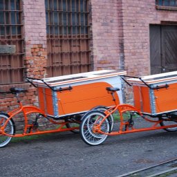 Alfa Welder - Pomarańczowy, dwuczęściowy rower cargo z białymi oponami na tle ceglanego budynku w Bąkowie.