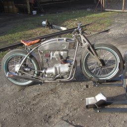 Alfa Welder - Surowy, metalowy motocykl typu custom na metalowym stojaku, z widocznymi spawami i elementami konstrukcyjnymi, na tle torów kolejowych i ziemi.