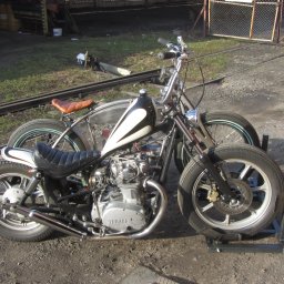 Alfa Welder - Dwa motocykle typu chopper zaparkowane obok siebie na szarym, żwirowym podłożu. Jeden czarny z białym bakiem, drugi metaliczny z brązowym siedzeniem. W tle widoczna brama z siatki i szyny kolejowe.
