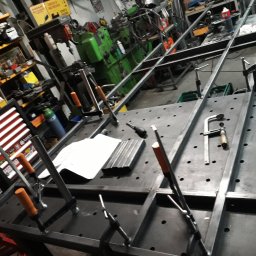 Alfa Welder - Metalowa konstrukcja spawana na stole warsztatowym z licznymi otworami, zabezpieczona ściskami stolarskimi, w tle widoczne narzędzia i maszyny warsztatowe.