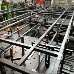 Alfa Welder - Konstrukcja z profili stalowych w trakcie spawania, zabezpieczona ściskami stolarskimi na perforowanym stole spawalniczym, widoczne narzędzia i elementy warsztatu w tle.