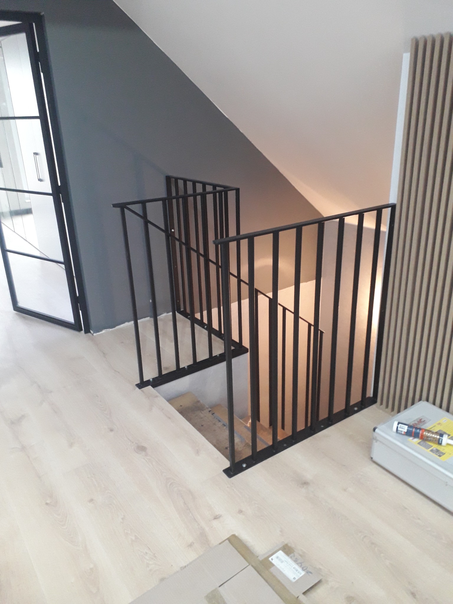 Nowoczesne schody z czarną metalową balustradą w minimalistycznym wnętrzu, widoczna podłoga z paneli w jasnym odcieniu drewna i szara ściana.