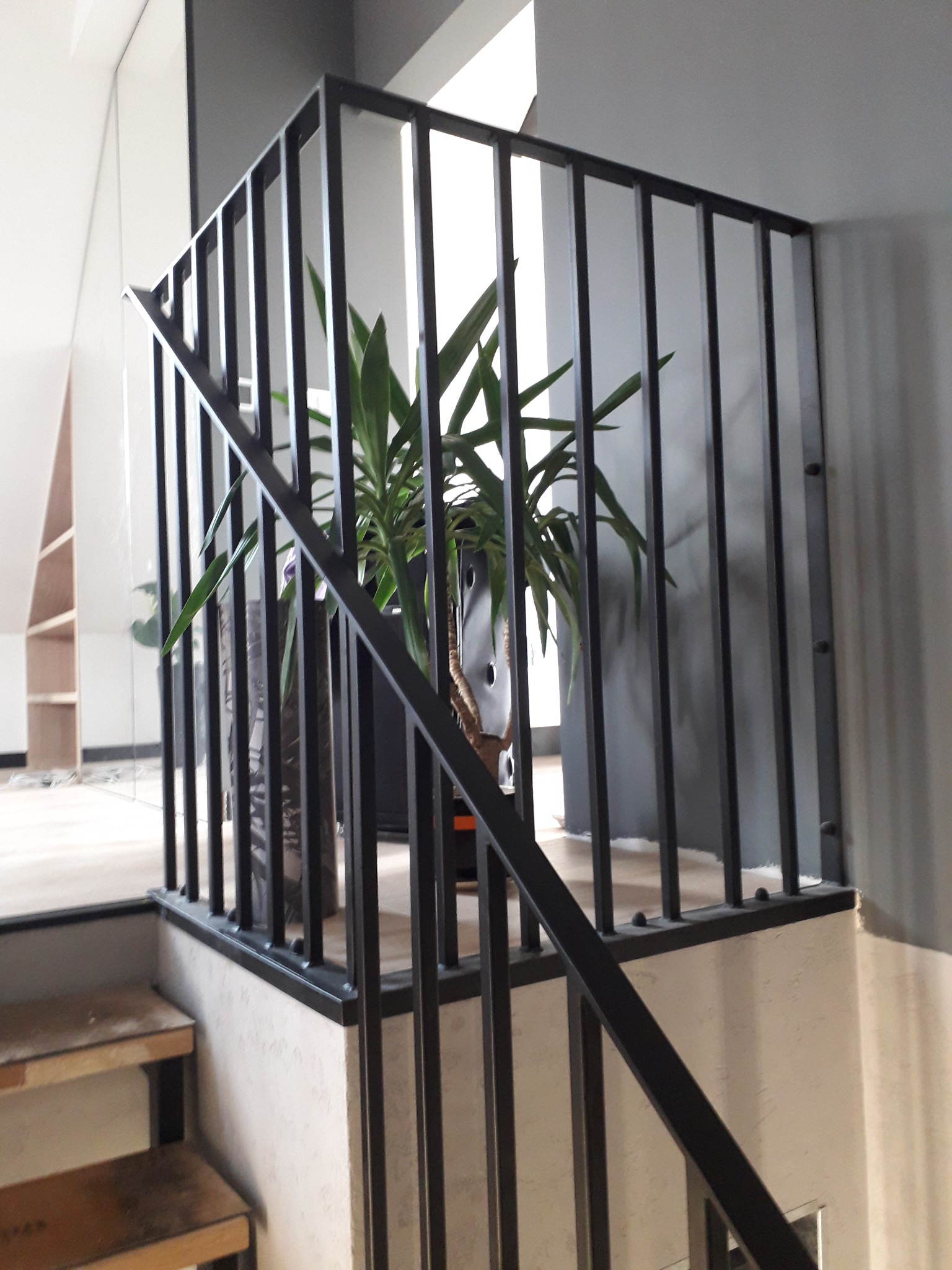 Nowoczesna, czarna metalowa balustrada schodowa z pionowymi szczebelkami, z doniczką z rośliną w tle, widok z dołu na schody w trakcie wykańczania.