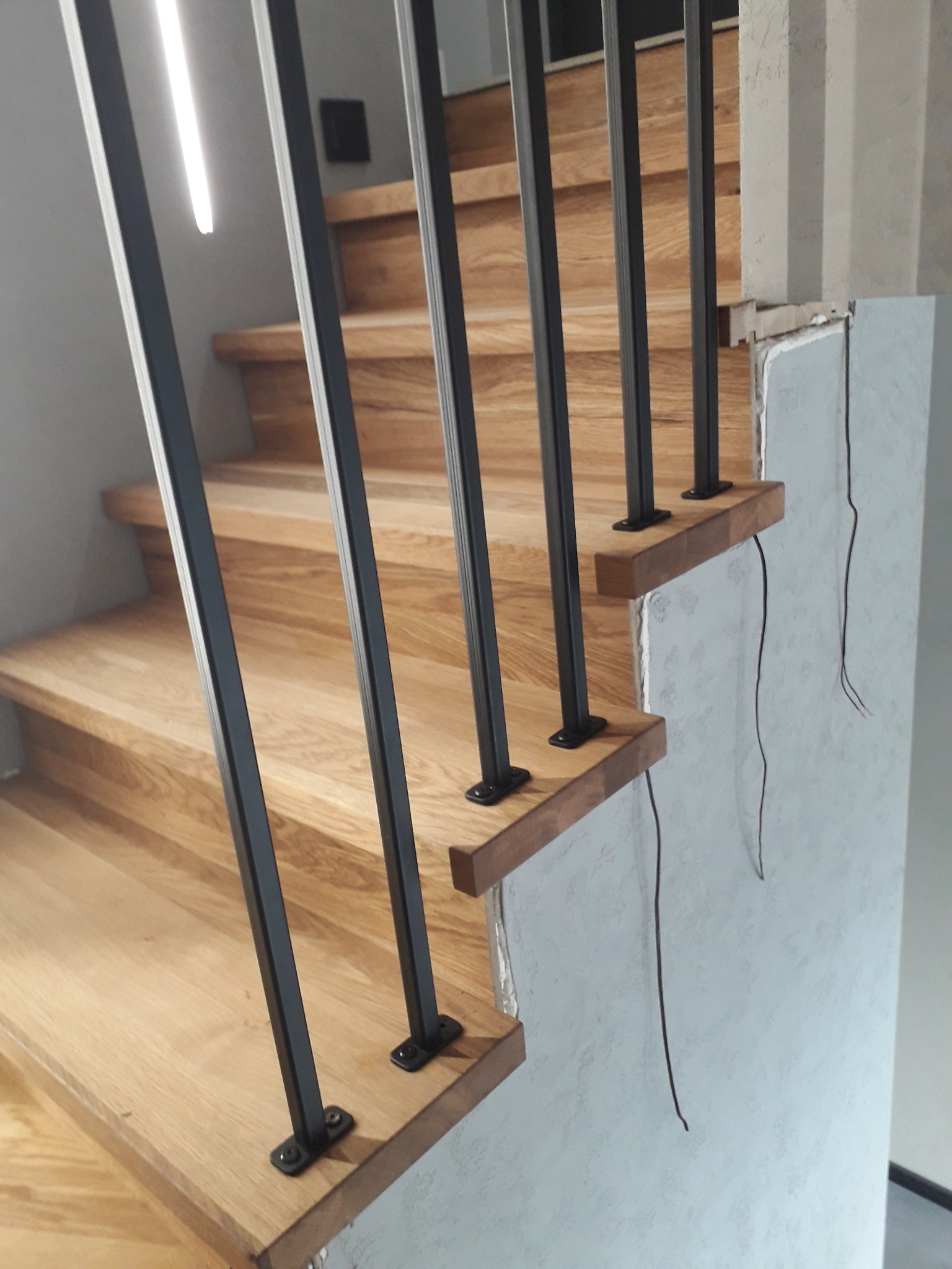 Fragment drewnianych schodów z czarną metalową balustradą, montowanych przy surowej ścianie z wystającymi przewodami elektrycznymi.