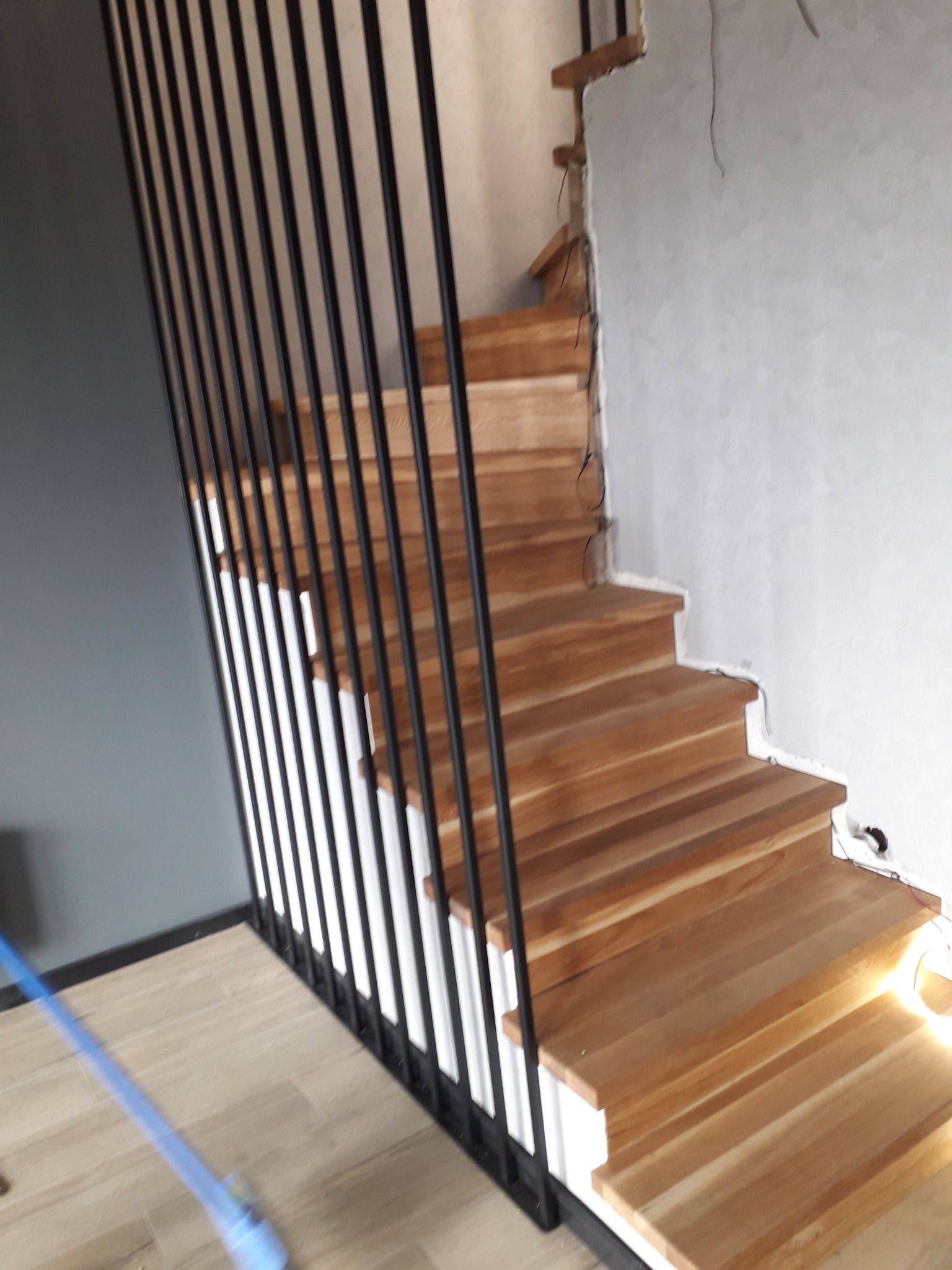 Drewniane schody z czarnymi, pionowymi balustradami, widoczne w trakcie montażu, z nie wykończonymi ścianami i wystającymi przewodami.