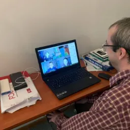 Mężczyzna w okularach uczestniczy w wideokonferencji na laptopie; na ekranie widoczni inni uczestnicy spotkania. Na biurku obok laptopa leżą paczki, drukarka i książki edukacyjne.