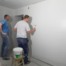 Dwóch mężczyzn w trakcie malowania ściany na biało za pomocą wałka. Widoczny kubeł z farbą na taborecie i narzędzia malarskie.