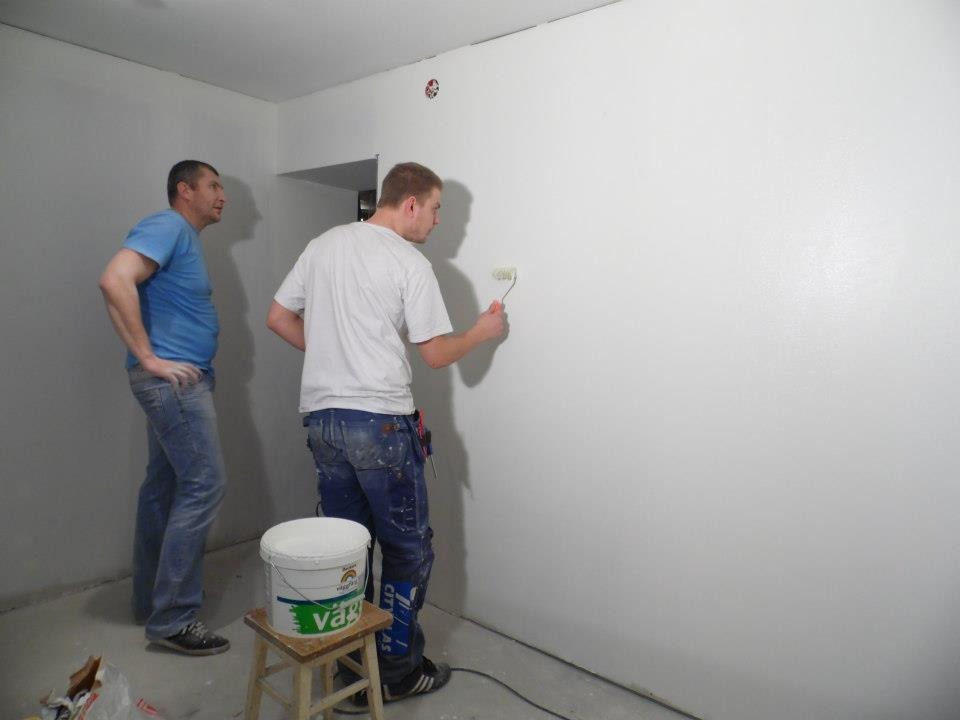 Dwóch mężczyzn w trakcie malowania ściany na biało za pomocą wałka. Widoczny kubeł z farbą na taborecie i narzędzia malarskie.