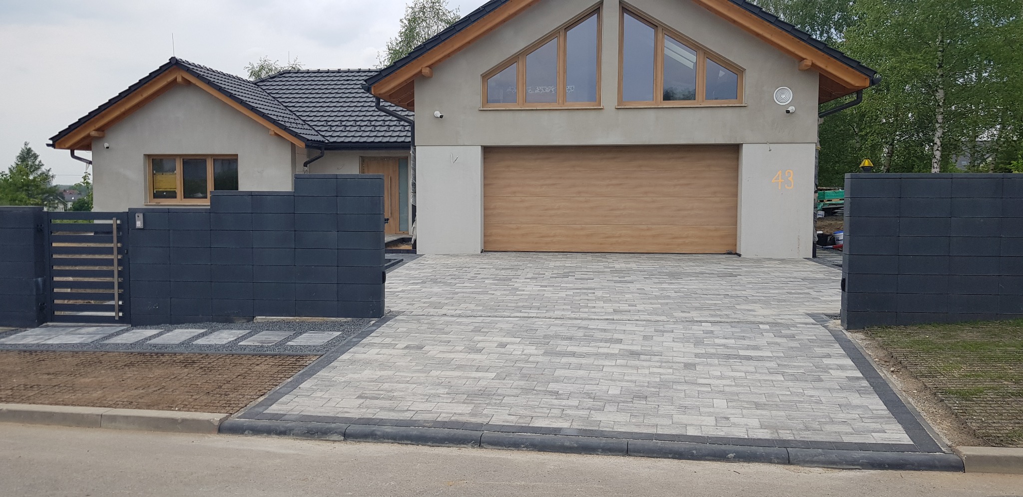 Ułożona szara kostka brukowa na podjeździe do nowoczesnego domu z drewnianą bramą garażową i grafitowym ogrodzeniem modułowym.