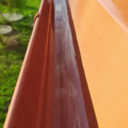 Wnętrze pomarańczowej rynny dachowej, częściowo umyte, z widoczną różnicą między czystą a zabrudzoną powierzchnią oraz z odbiciem nieba i zieleni.