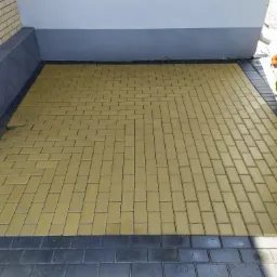 Świeżo ułożona, jasna kostka brukowa w jodełkę, obramowana ciemniejszymi kostkami, przylegająca do ściany budynku z rynną i zielonym krzewem.