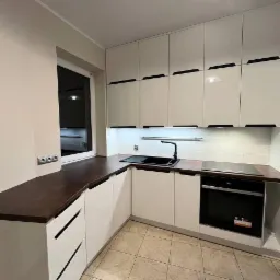 Nowoczesna kuchnia z białymi, lakierowanymi frontami, ciemnym blatem drewnianym i czarnymi uchwytami, oświetlenie podszafkowe LED, płytki podłogowe w odcieniach beżu.