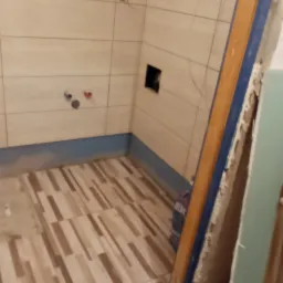 Wnętrze łazienki w trakcie remontu: ułożone płytki podłogowe w odcieniach beżu i brązu, częściowo wykończone ściany z jasnymi płytkami, widoczne narzędzia i materiały budowlane na podłodze, w tym...