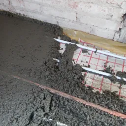 Wylewka betonowa pokrywająca rury ogrzewania podłogowego na folii z siatką, widoczny fragment ceglanej ściany w tle.