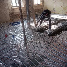 Montaż ogrzewania podłogowego w surowym pomieszczeniu z odsłoniętą cegłą, widoczne rury grzewcze na siatce, pracownik w trakcie mocowania, puszki po napojach na podłodze.