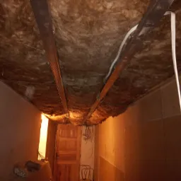 Poddasze w trakcie ocieplania, widoczne metalowe profile, izolacja z wełny mineralnej oraz wystające przewody elektryczne, w tle drewniane drzwi i fragment ściany.