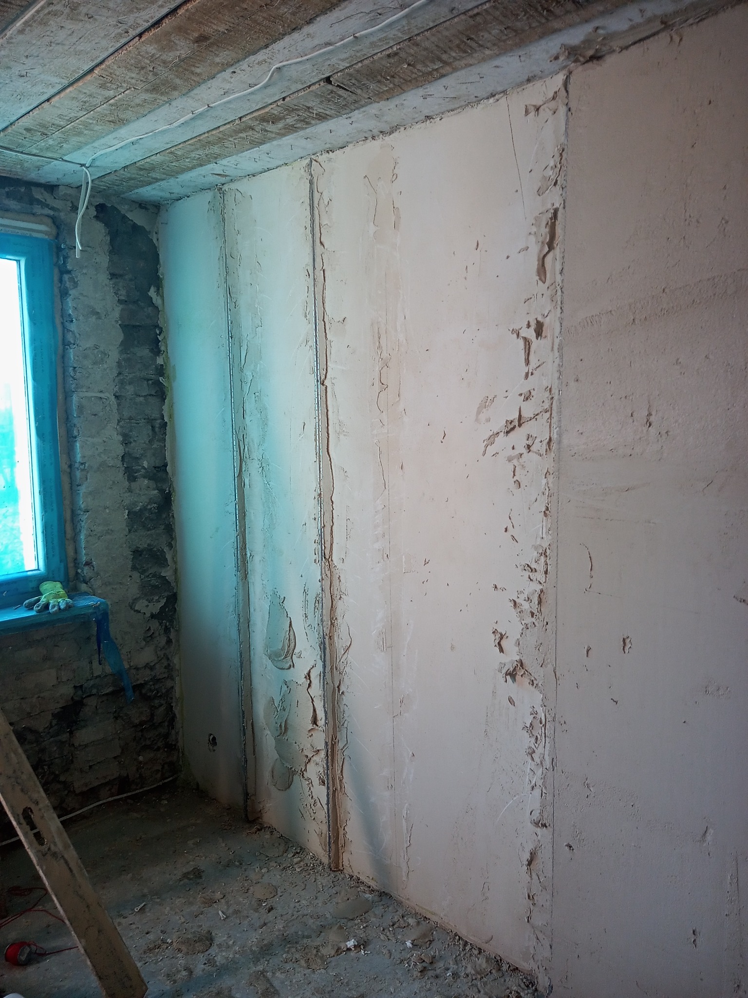 Wnętrze w trakcie remontu: przygotowanie ścian pod płyty gipsowo-kartonowe, widoczne profile pionowe i częściowo nałożona masa szpachlowa, surowe drewniane belki stropowe, okno z nieotynkowanym...