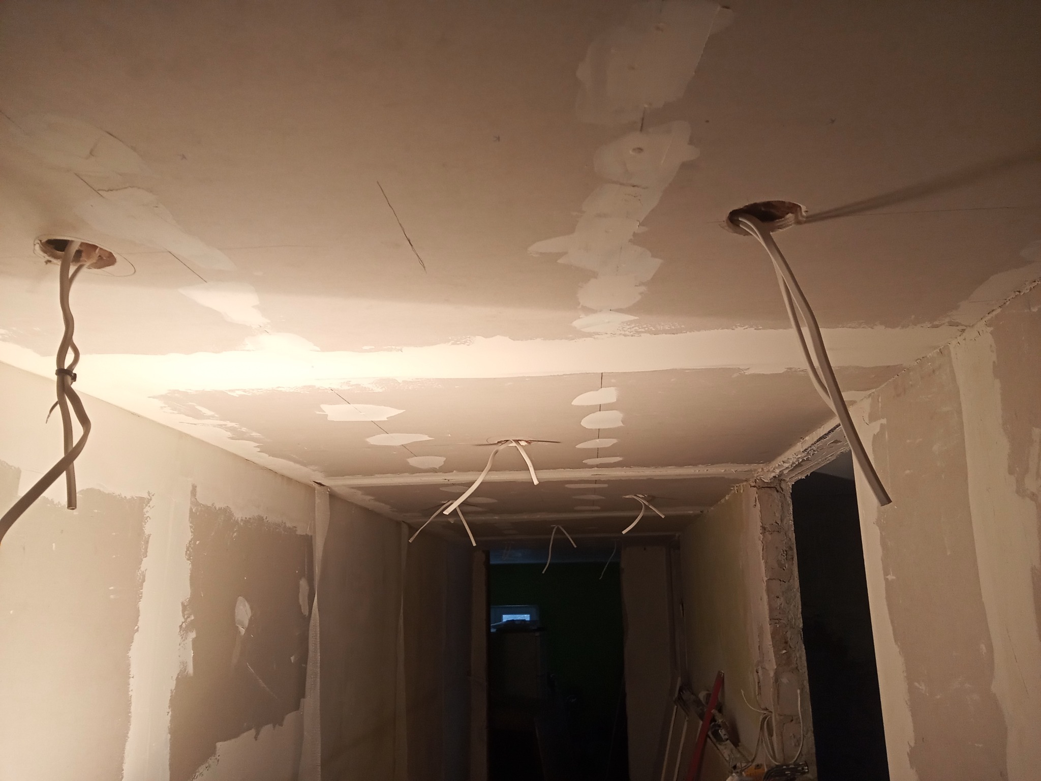 Wnętrze w trakcie remontu: surowy sufit z widocznymi przewodami elektrycznymi wystającymi z otworów, ściany w stanie surowym, częściowo pokryte gładzią, widoczne ślady szpachlowania na suficie...