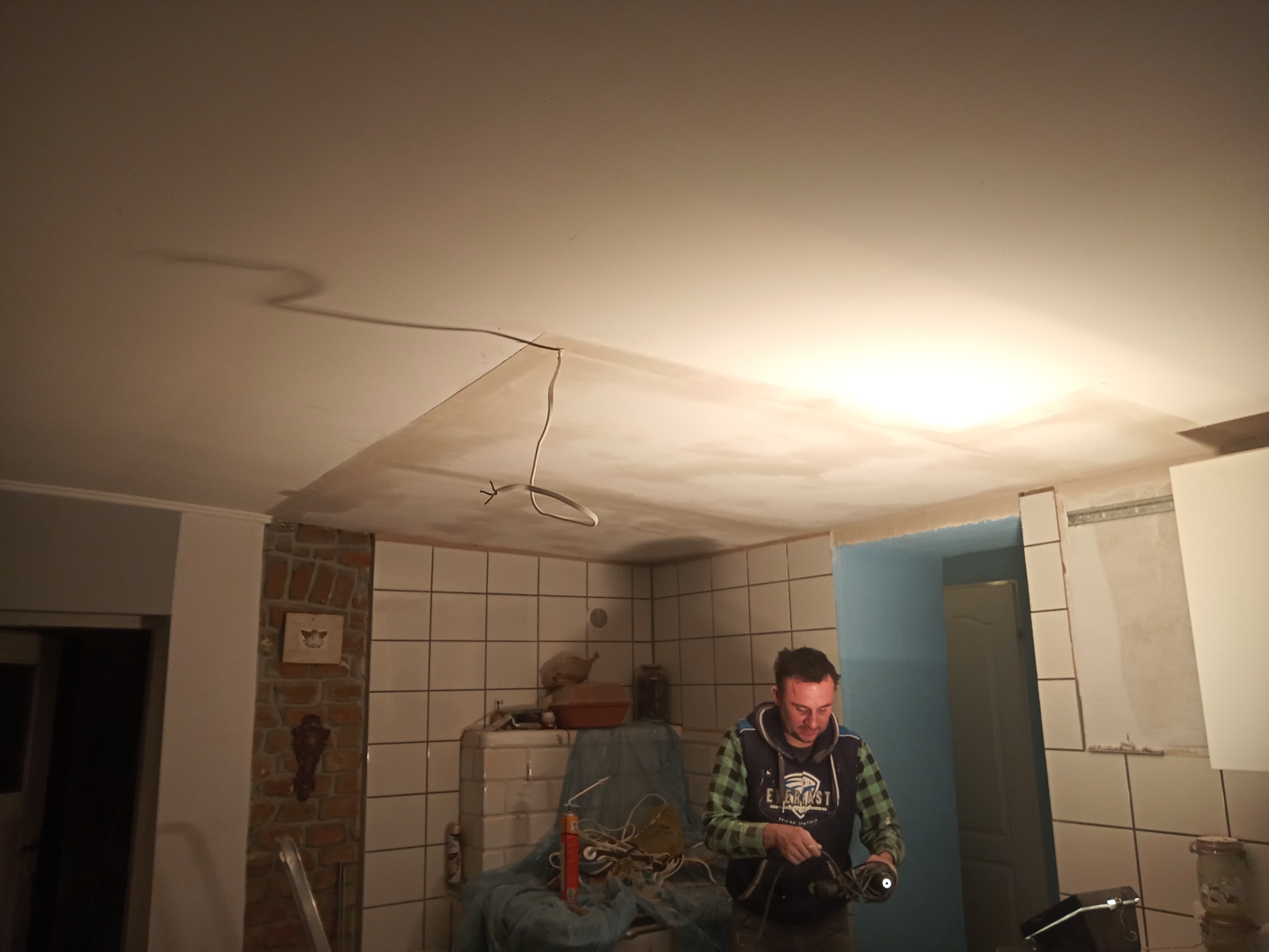 Mężczyzna w trakcie prac remontowych w łazience z widoczną instalacją elektryczną na suficie i częściowo położonymi płytkami na ścianach.