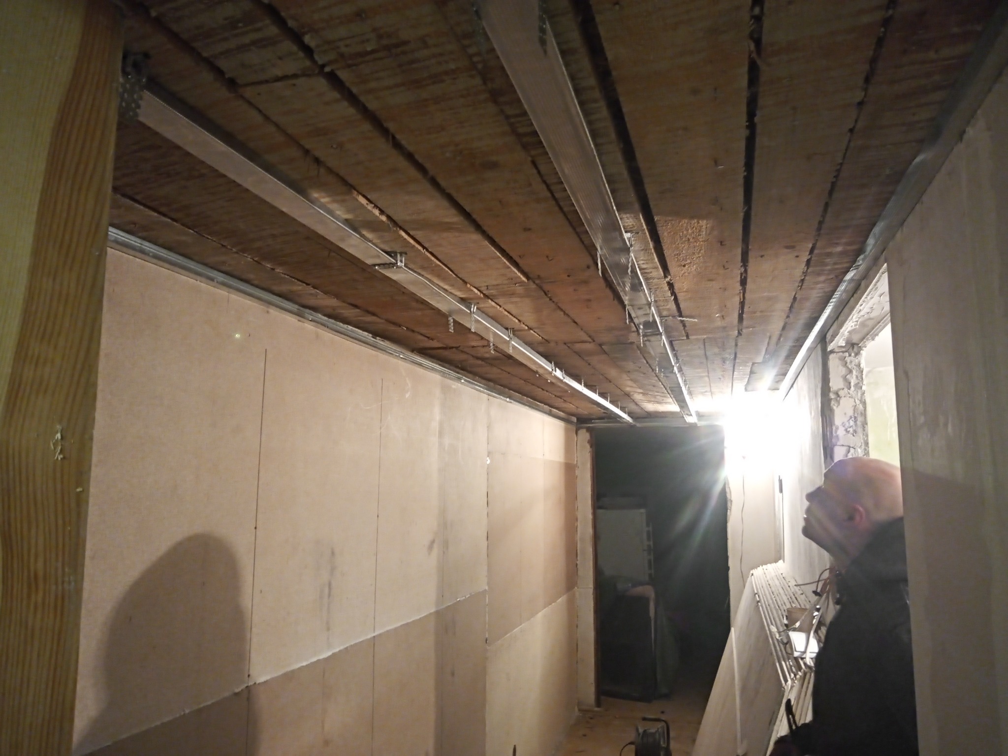 Montaż metalowego stelażu pod płyty karton-gips na drewnianym stropie, widoczny fragment ściany z zamontowanymi płytami oraz sylwetka mężczyzny w tle.