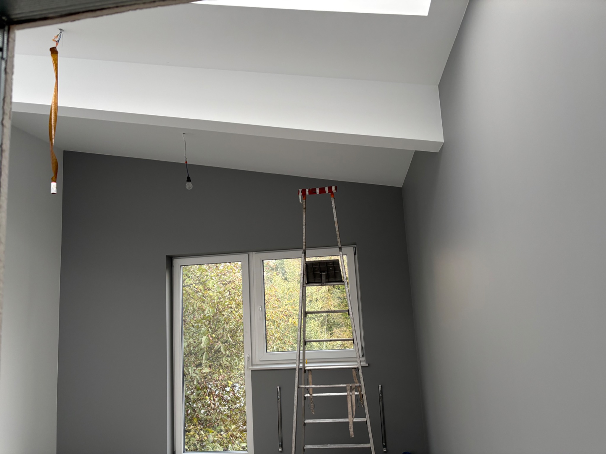 Wnętrze pokoju po malowaniu na szaro z białą listwą pod sufitem, drabina oparta o okno, kabel zwisający z sufitu. Widok na drzewa za oknem.