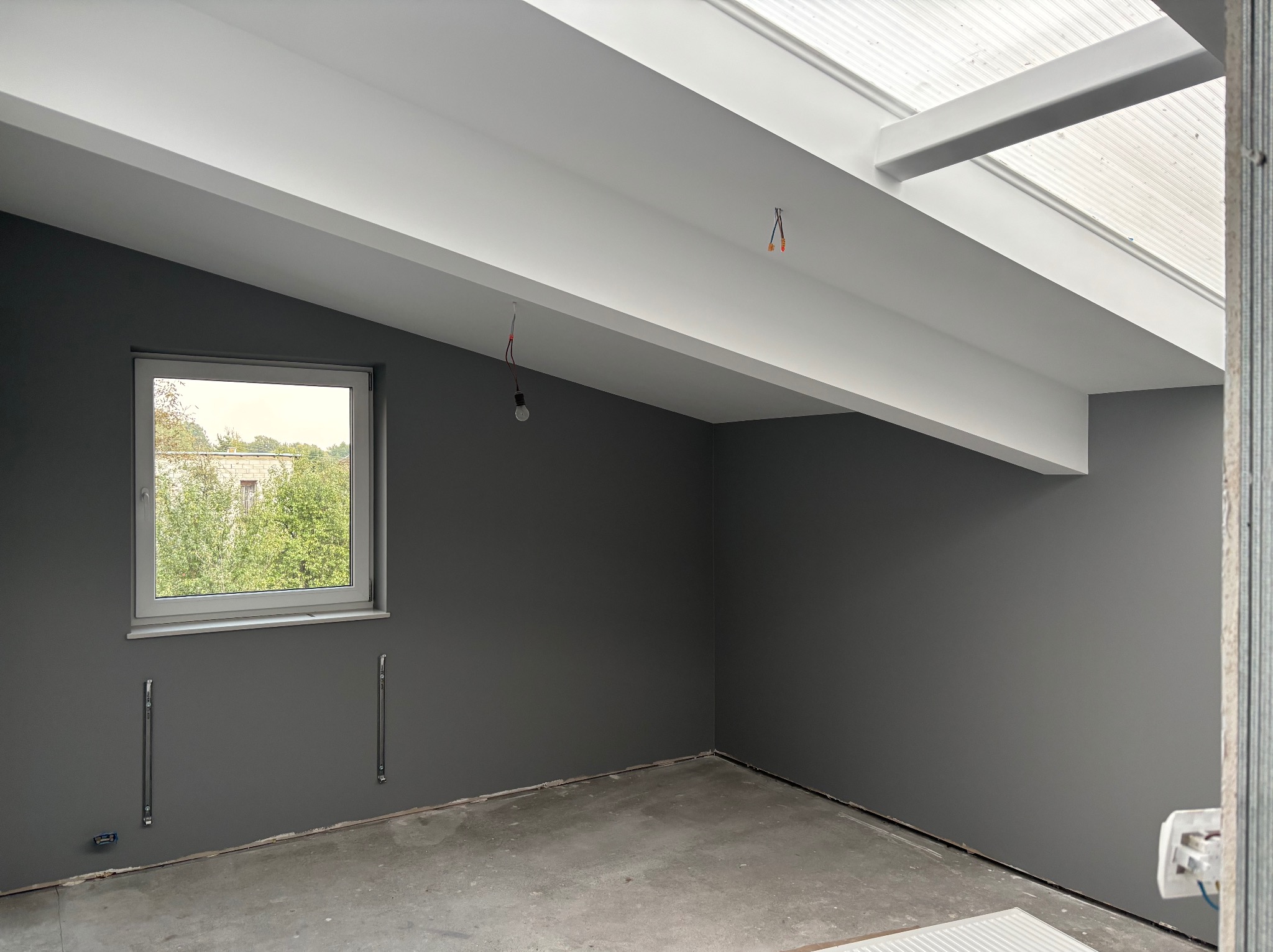 Szare ściany w pokoju na poddaszu z oknem i białymi belkami stropowymi, widoczne przewody elektryczne i elementy montażowe, surowy beton na podłodze.