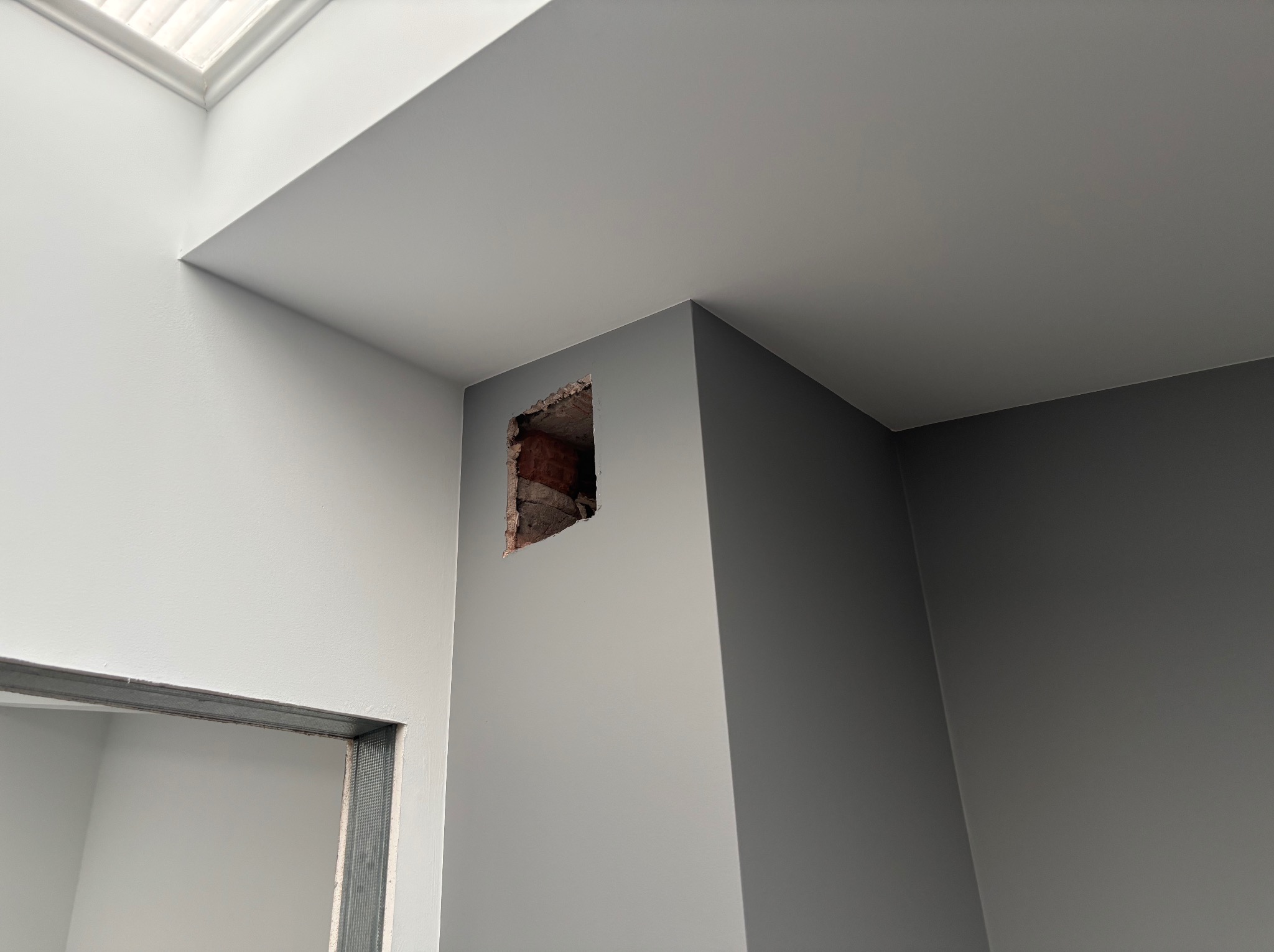 Wnętrze po malowaniu ścian na szaro i biało. Widoczne otwory w ścianie po instalacjach i fragment metalowej ramy drzwiowej. Oświetlenie naturalne z okna dachowego.