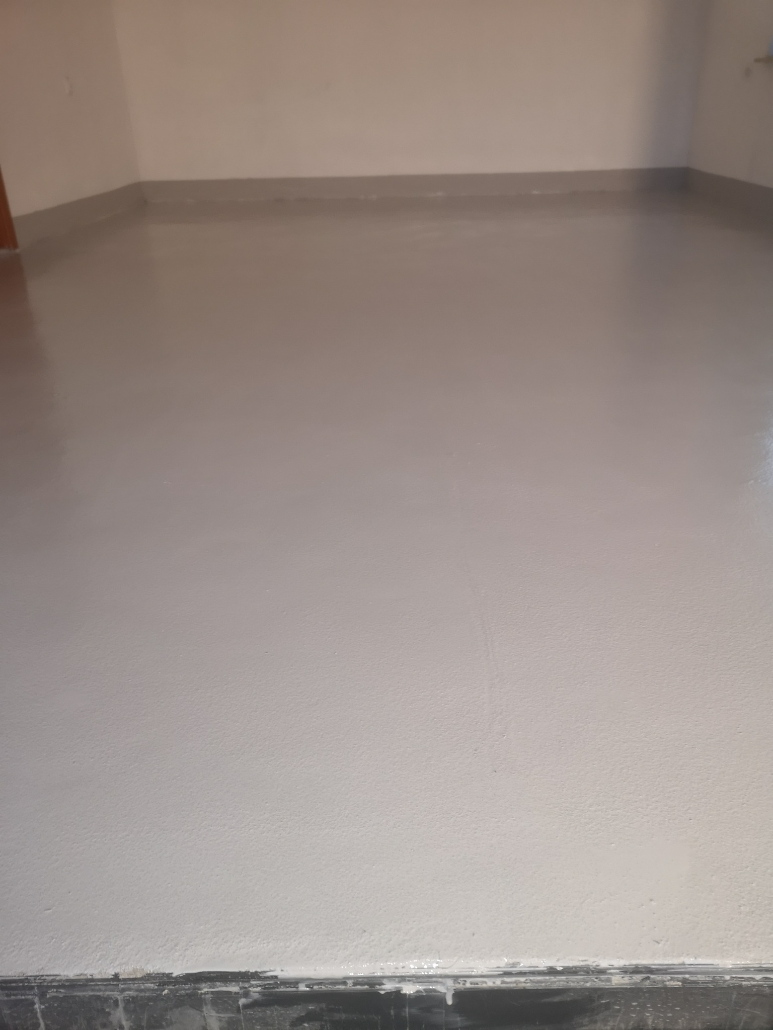 Gładka, jasnoszara posadzka w pomieszczeniu z listwami przypodłogowymi, oświetlona naturalnym światłem, ukazująca jednolitą powierzchnię.