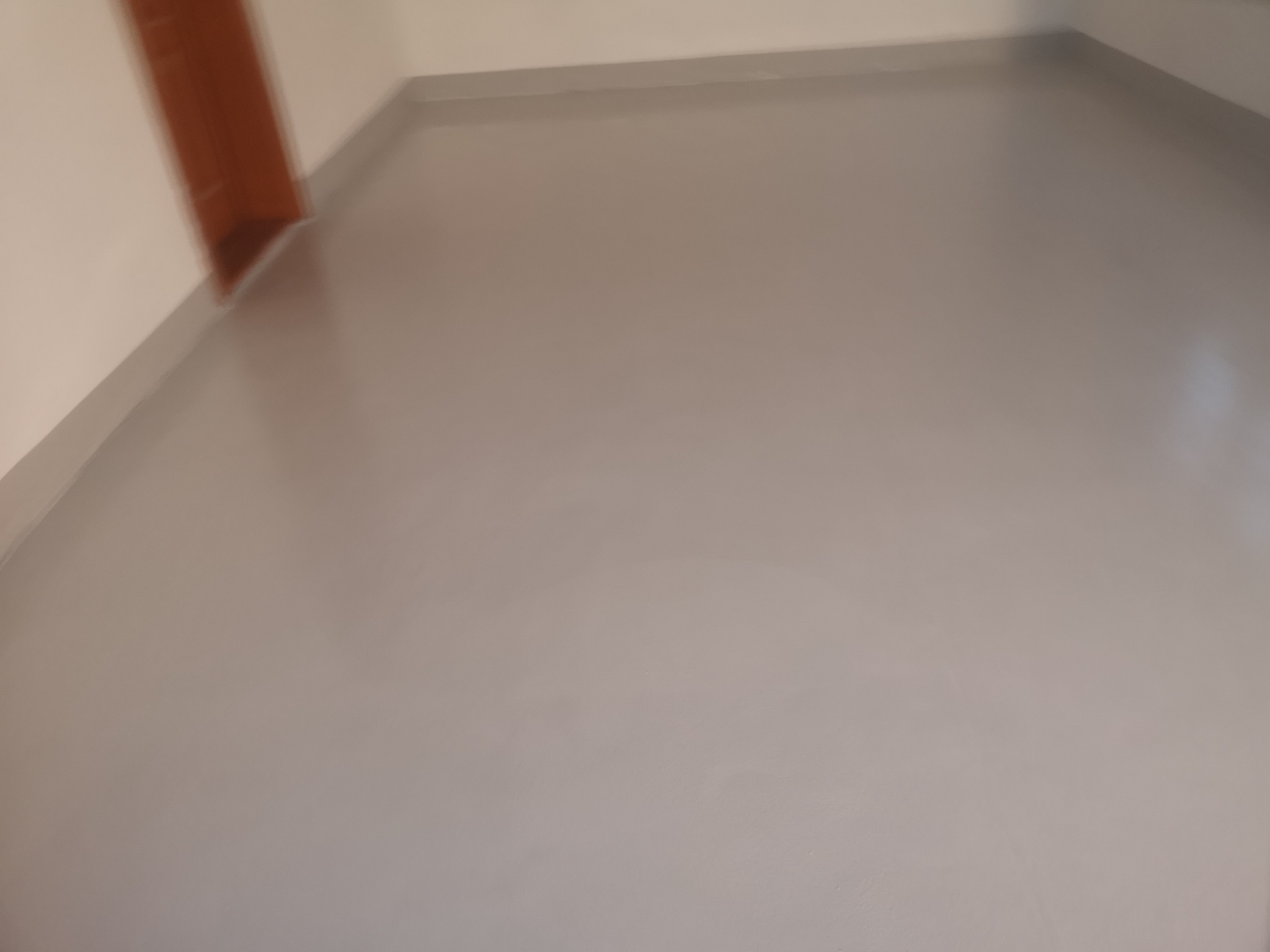 Jasnoszara, gładka powierzchnia podłogi z widocznym łączeniem ze ścianą, przypominająca wylewkę żywiczną.