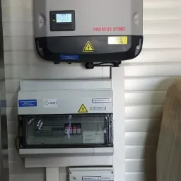 Szary falownik Fronius Symo zamontowany na ścianie z panelami wraz z rozdzielnicami PV-AC i PV-DC, widoczne ż&oacute;łte naklejki ostrzegawcze.