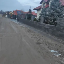 Nowo postawiony szary, betonowy mur ogrodzeniowy z filarami, widok z zabłoconej drogi, w tle domy jednorodzinne i pracownik na skuterze.