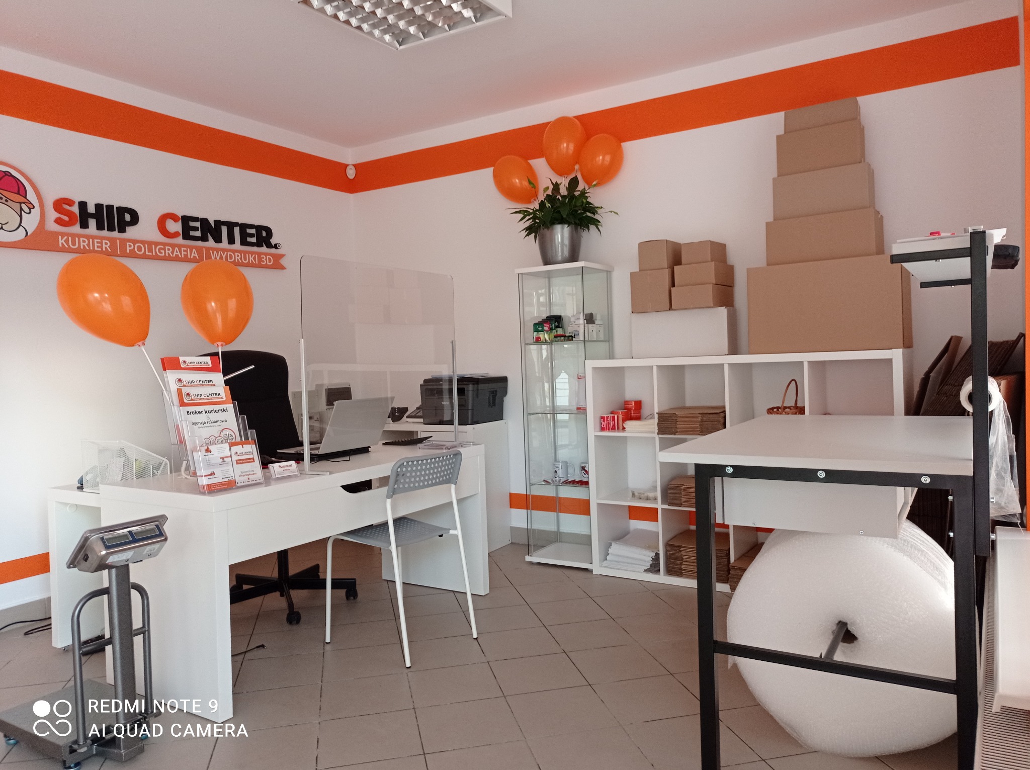 Wnętrze punktu kurierskiego: biurko z laptopem i wagą, za szybą z pleksi, pomarańczowe balony i logo Ship Center na ścianie, kartony w tle.