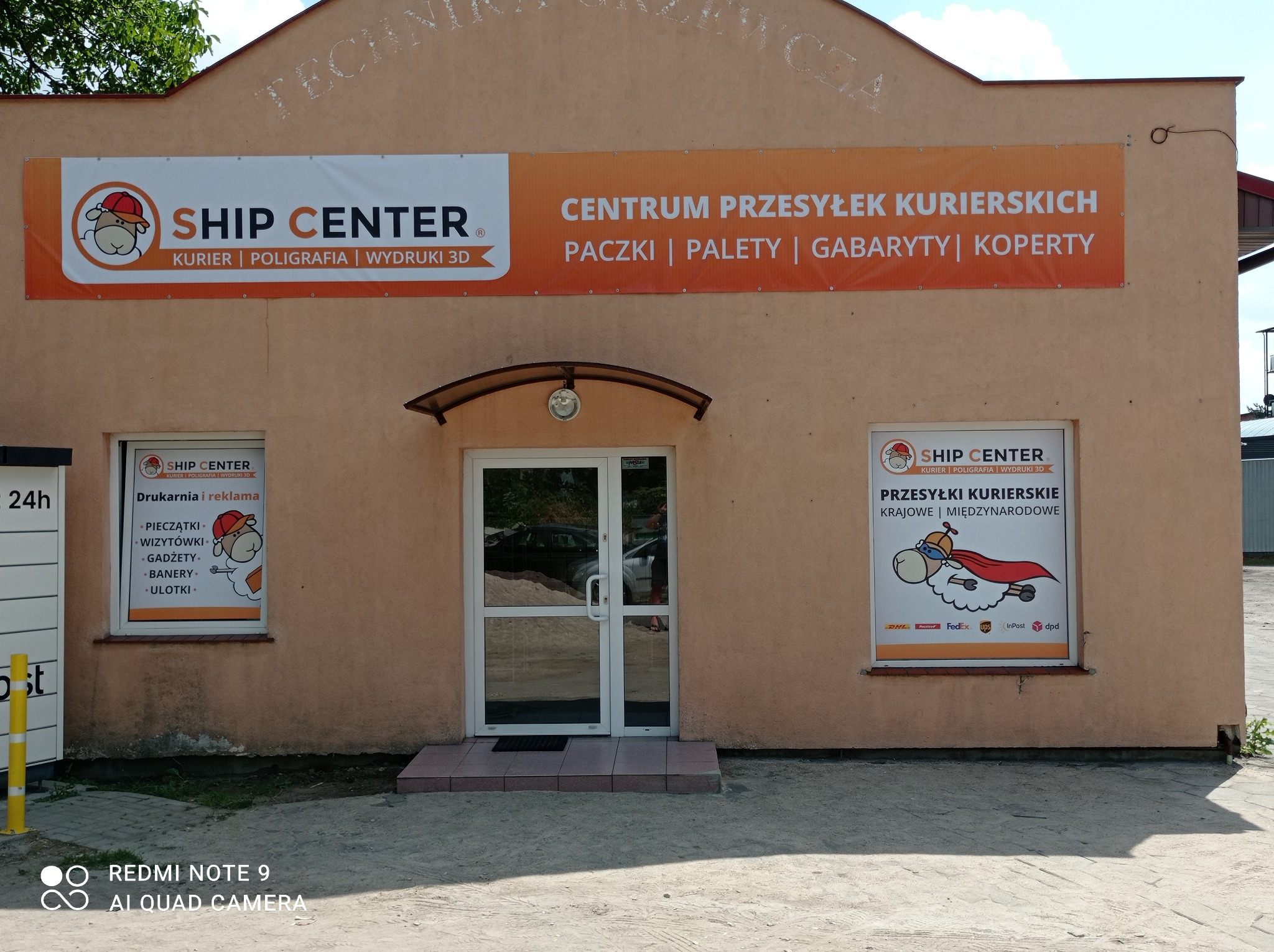 Fasada budynku firmy kurierskiej Ship Center z dużym banerem reklamowym nad wejściem, informującym o ofercie: przesyłki kurierskie, paczki, palety, gabaryty, koperty. Po bokach okna z plakatami...