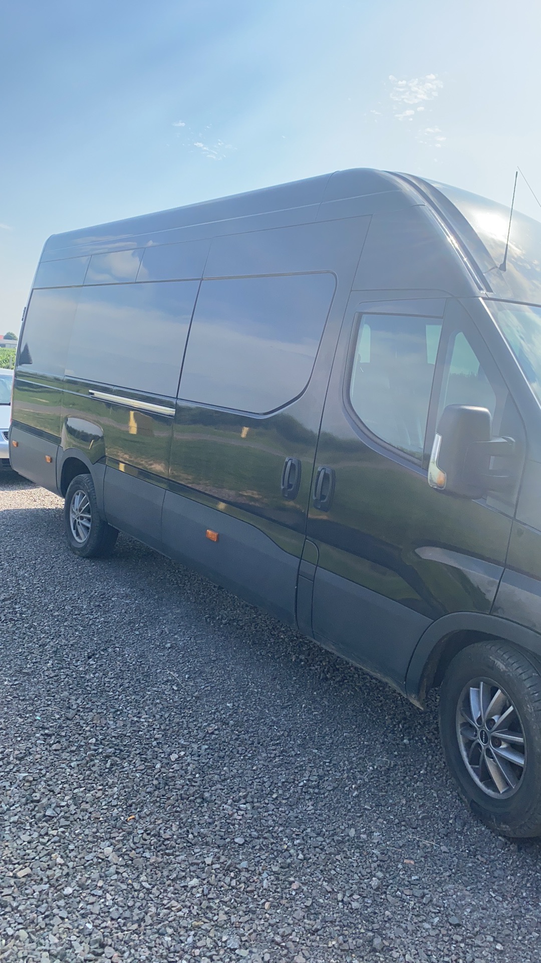 Czarny, przedłużony bus transportowy na żwirowym podłożu, widoczny z boku, z polami w tle pod błękitnym niebem.