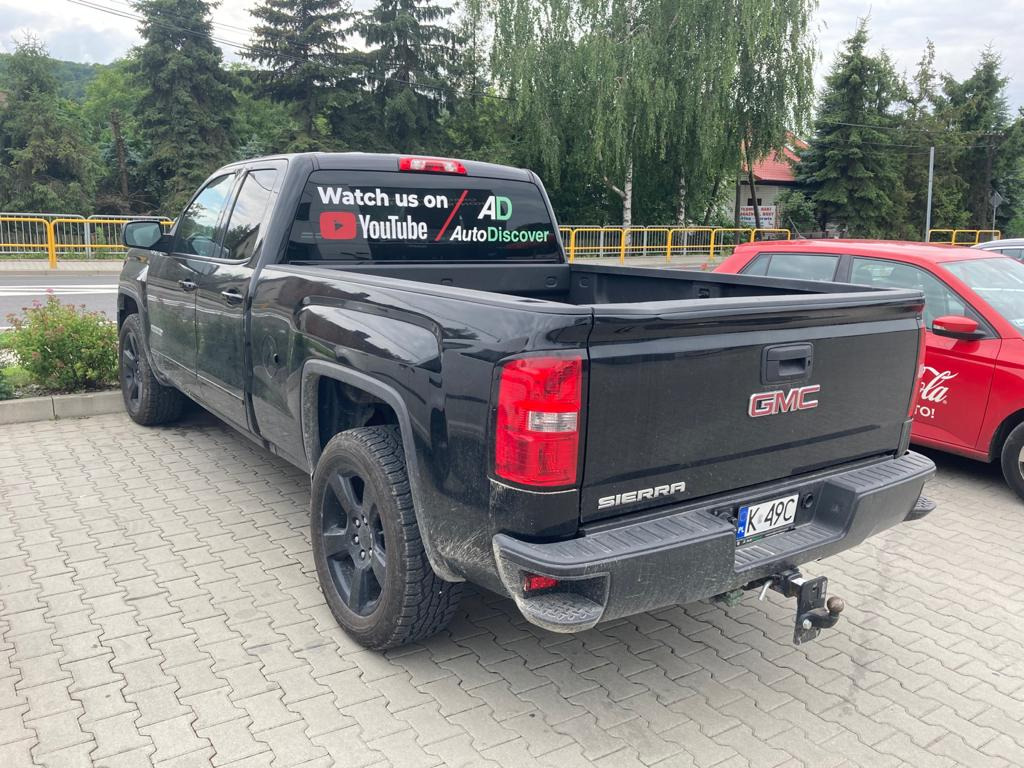 Czarny pick-up GMC Sierra z przyczepą holowniczą, zaparkowany na brukowanej powierzchni, z naklejkami YouTube i AutoDiscover na tylnej szybie.