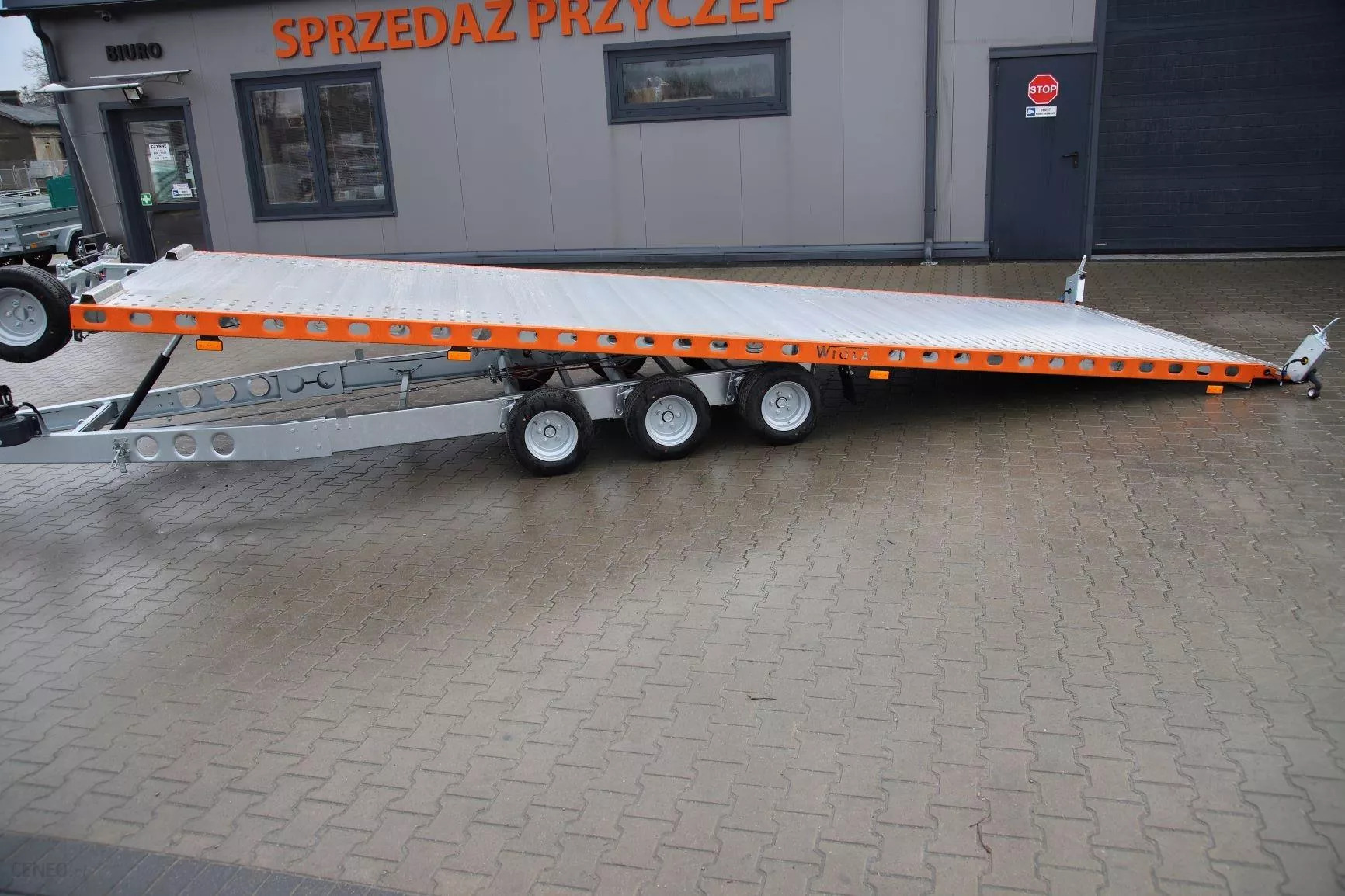 Pusta, trójosiowa laweta samochodowa z pomarańczowymi obramowaniami, stojąca na kostce brukowej przed budynkiem z napisem 'Sprzedaż Przyczep'.