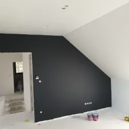 Pokój na poddaszu w trakcie remontu: pomalowana na czarno ściana szczytowa, białe ściany boczne i sufit, widoczne puszki farby Śnieżka oraz niedokończone schody w przejściu do innego pomieszczenia.