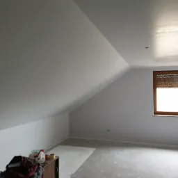 Poddasze po szpachlowaniu i malowaniu na biało, widoczne gładkie ściany ze skosem, okno z brązowymi roletami oraz prowizoryczne zabezpieczenie podłogi folią malarską. Na pierwszym planie fragment...