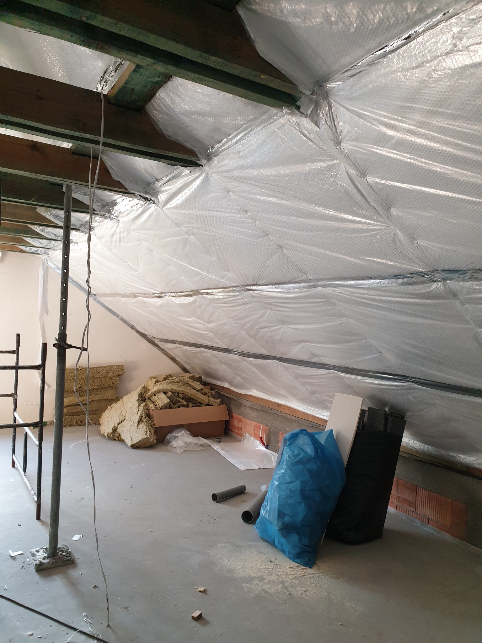 Poddasze w trakcie ocieplania, widoczna folia paroizolacyjna na skośnych ścianach, rusztowanie, wełna mineralna w kartonie, oraz worki na odpady budowlane.