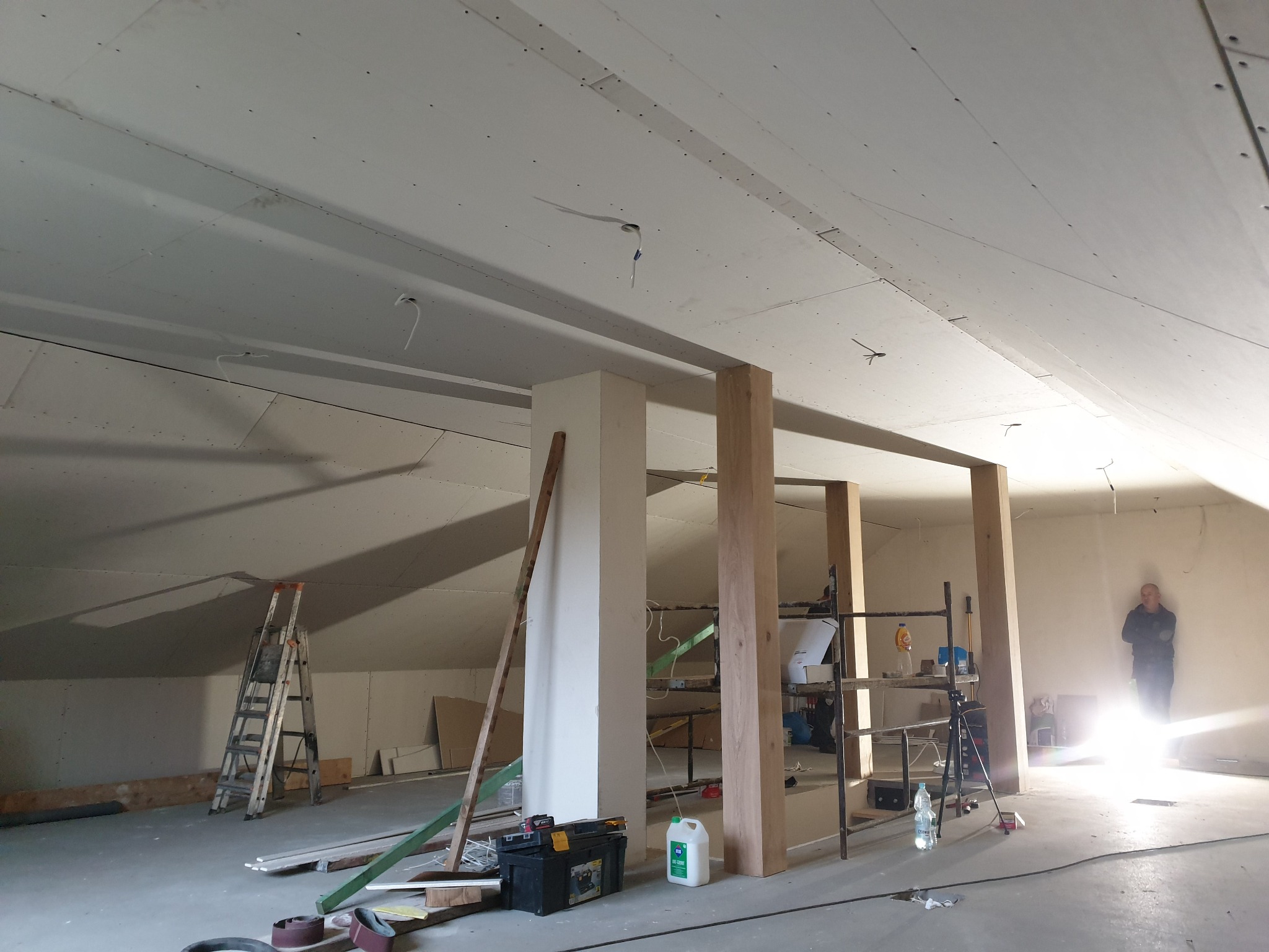 Poddasze w Barlinku w trakcie remontu, widoczne płyty karton gips na suficie, konstrukcja szkieletowa z drewna, drabina i narzędzia, sylwetka osoby w tle przy oknie.