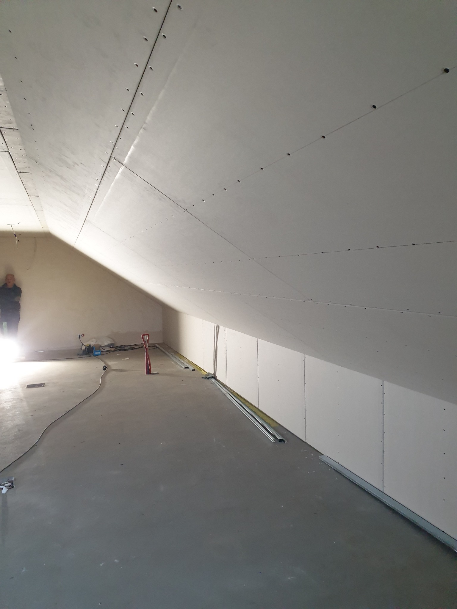 Montaż płyt kartonowo-gipsowych na skośnym poddaszu, widoczne profile i narzędzia, osoba w tle. Świeżo położona wylewka podłogowa.