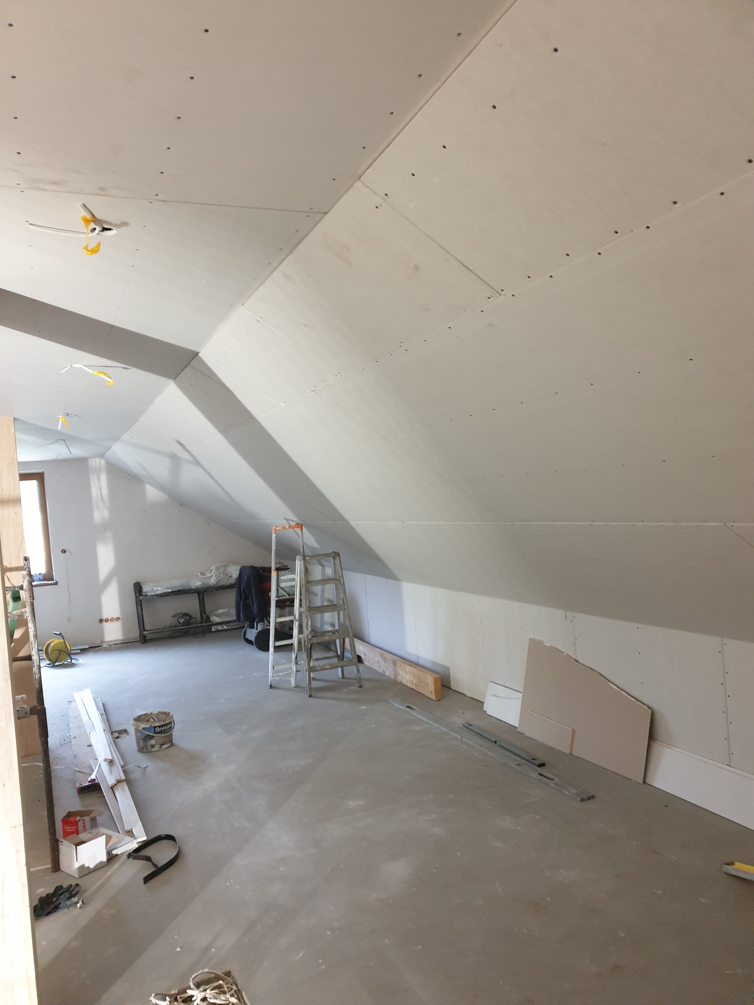 Poddasze w trakcie wykańczania płytami kartonowo-gipsowymi, widoczne łączenia płyt na skosach, drabina, narzędzia i materiały budowlane porozrzucane na podłodze.