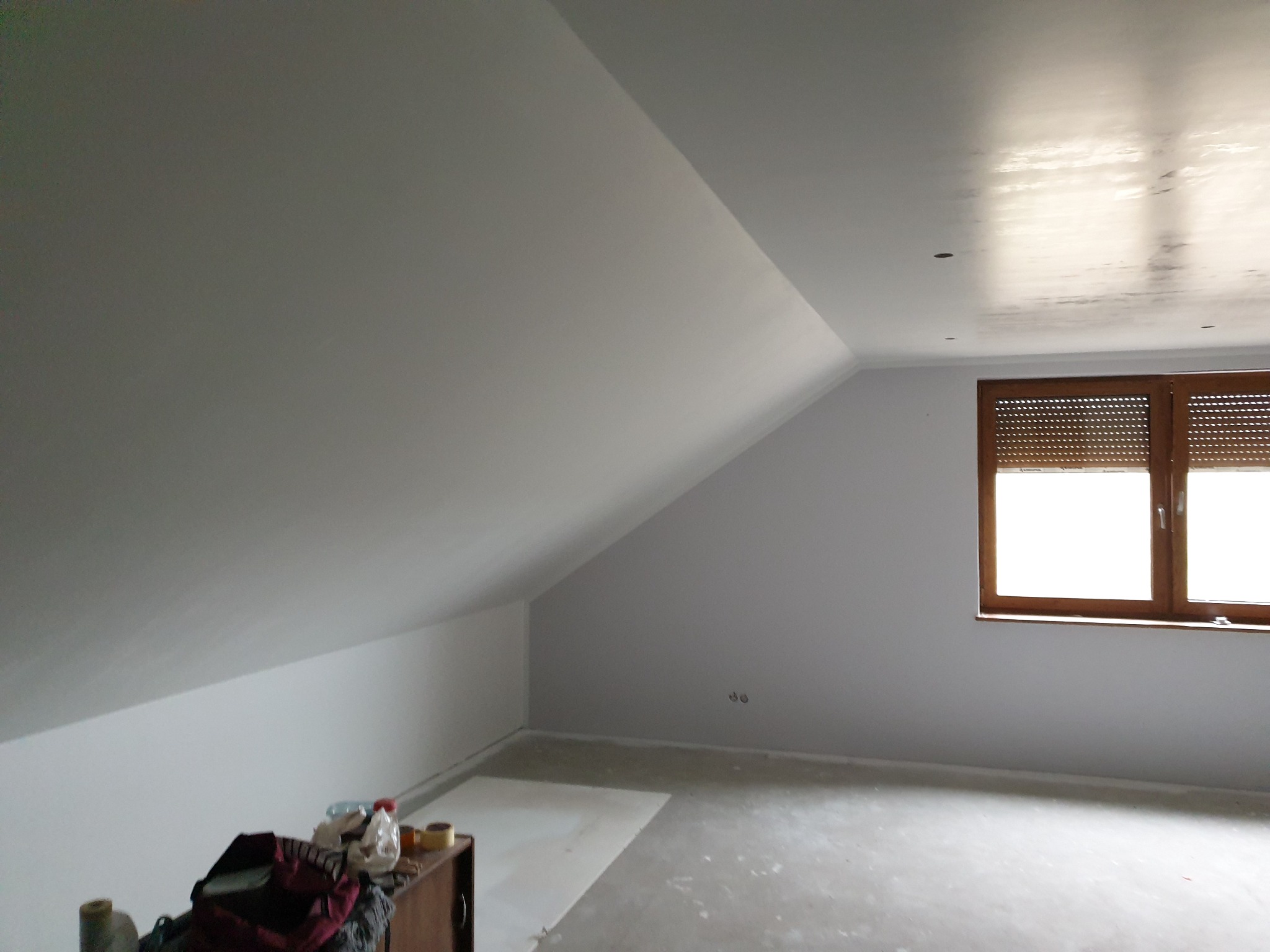 Poddasze po szpachlowaniu i malowaniu na biało, widoczne gładkie ściany ze skosem, okno z brązowymi roletami oraz prowizoryczne zabezpieczenie podłogi folią malarską. Na pierwszym planie fragment...