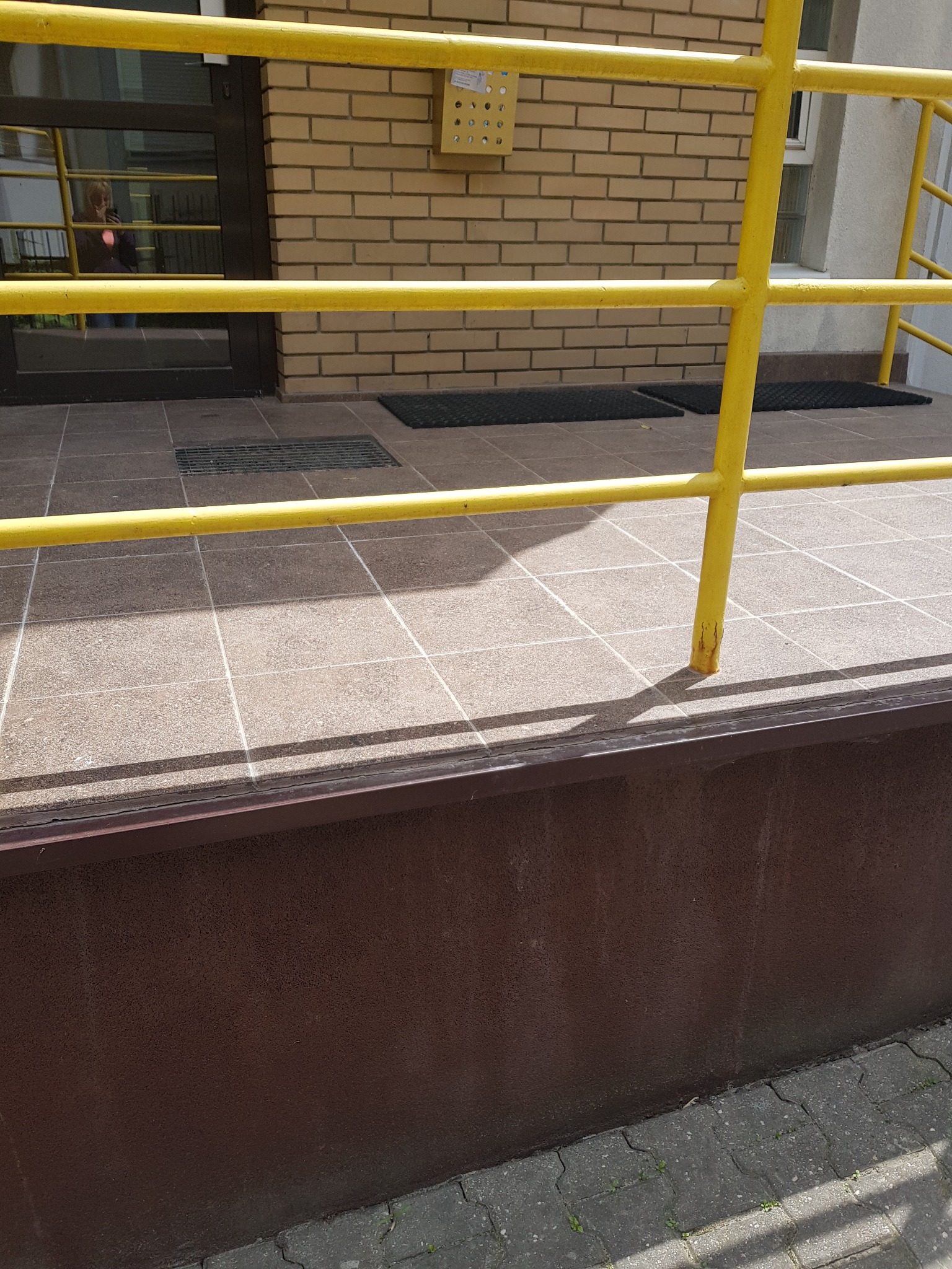 Wejście do budynku z żółtą barierką, widoczne wycieraczki i ceglana ściana