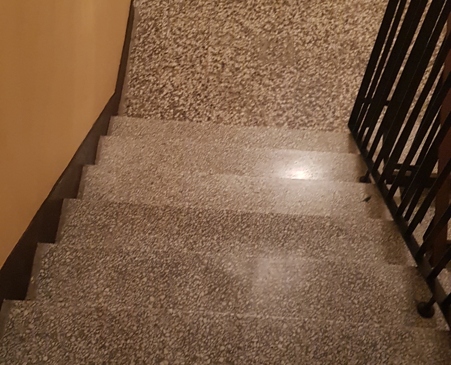 Schody z lastryko w budynku, widok z góry, z metalową barierką po prawej stronie i żółtą ścianą po lewej.