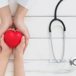 Dłonie dorosłego i dziecka trzymają czerwone serce, obok leży stetoskop na białym drewnianym tle.