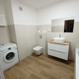 Nowoczesna łazienka po remoncie: biała umywalka nablatowa na szafce z drewnianym blatem, toaleta podwieszana, pralka Bosch i płytki imitujące drewno na podłodze.