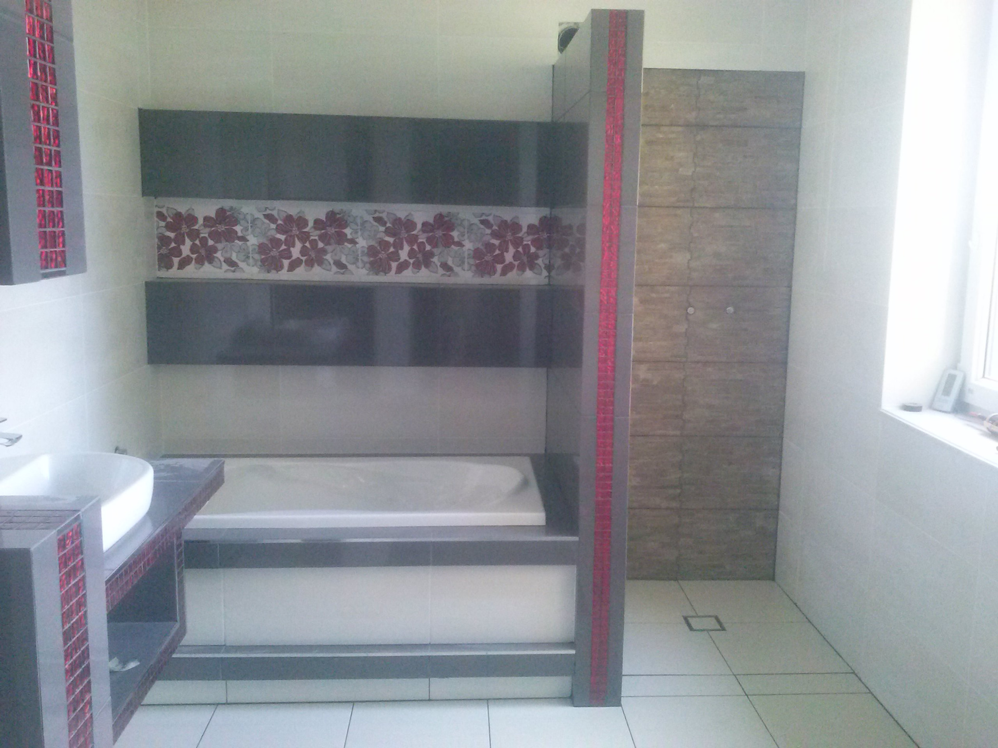 Nowoczesna łazienka z wanną obudowaną szarymi płytkami z motywem kwiatowym, umywalką z szafką zdobioną mozaiką, oraz kabiną prysznicową z płytek imitujących kamień.