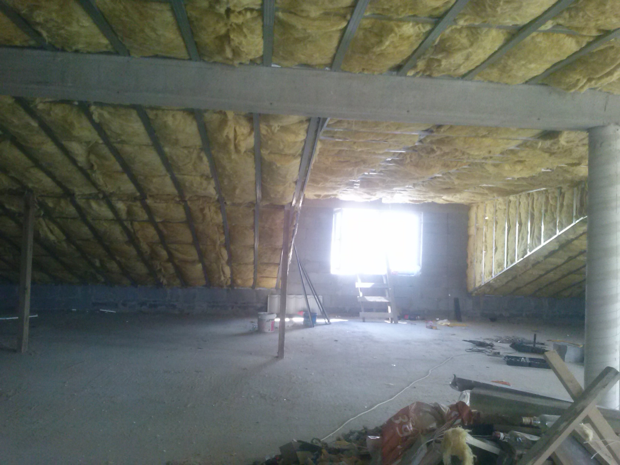 Poddasze w trakcie izolacji termicznej, widoczne wełna mineralna między krokwiami, surowe ściany i podłoga, drabina oparta o ścianę z oknem.